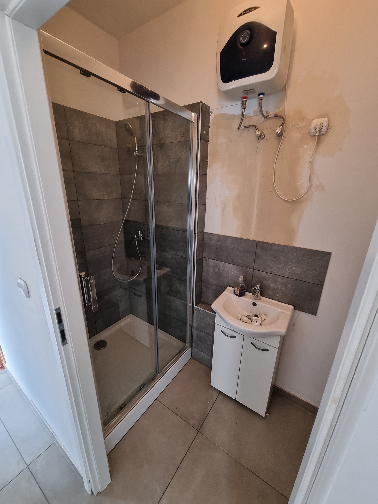 Łazienka w trakcie prac: kabina prysznicowa z ciemnymi płytkami, biała szafka z umywalką, bojler elektryczny na ścianie z widocznymi śladami po instalacji.