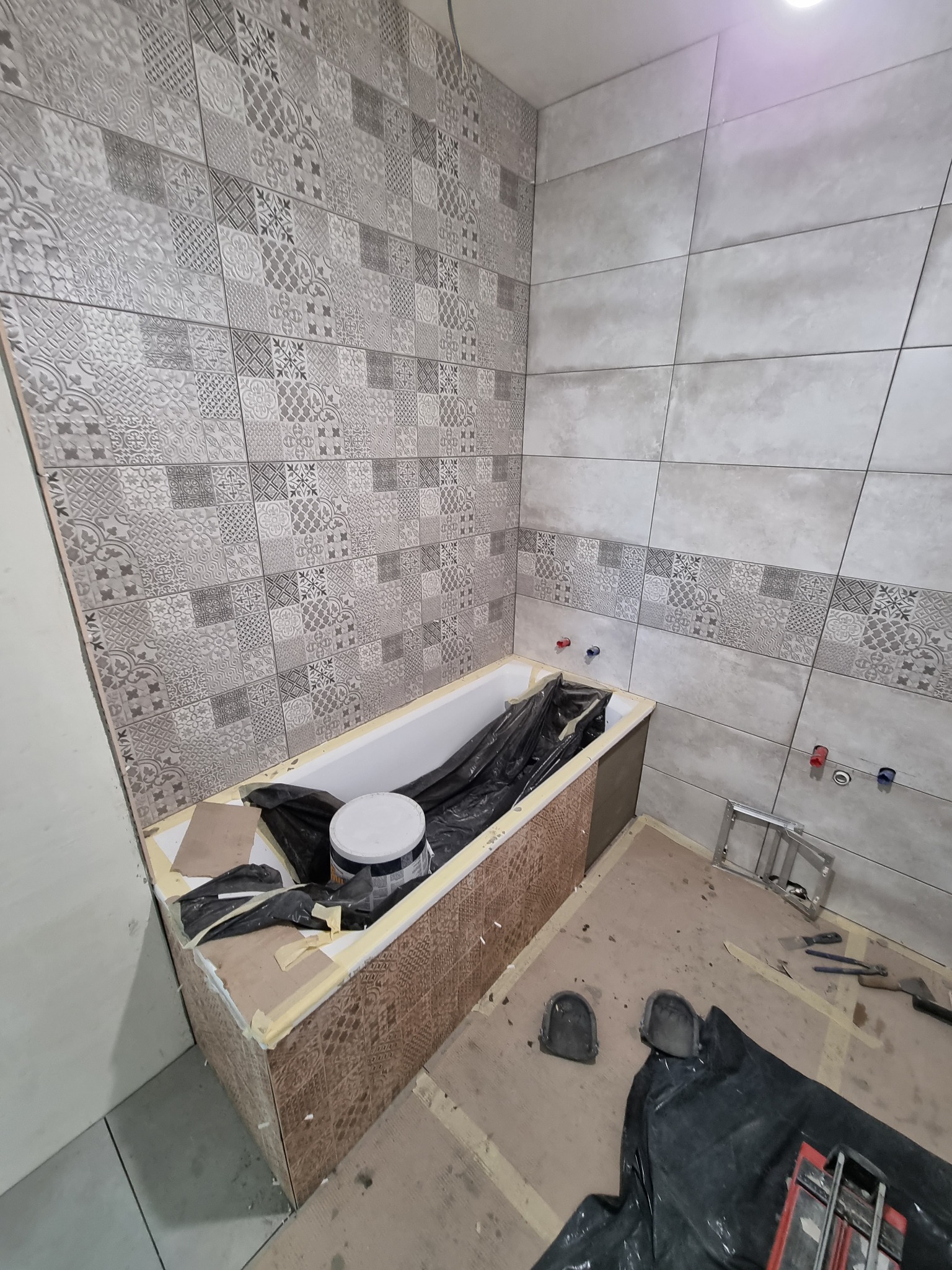 Łazienka w trakcie prac: obudowa wanny wyłożona brązowymi płytkami, ściany częściowo z szarymi płytkami o wzorach patchwork, częściowo z dużych szarych płyt, podłoga zabezpieczona folią, widoczne...