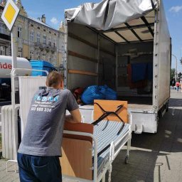 Greg-Trans Grzegorz Śliwa - Mężczyzna w szarej koszulce z logo firmy przeprowadzkowej opiera się o wózek transportowy z łóżkiem szpitalnym na tle otwartej paki samochodu dostawczego, widoczny budynek i rowerzysta.
