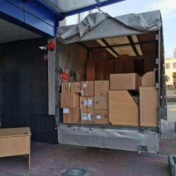 Greg-Trans Grzegorz Śliwa - Otwarta paka ciężarówki wypełniona kartonami i meblami, częściowo przykryta szarą plandeką, zaparkowana na brukowanym placu z biurkiem obok, widoczny budynek w tle.