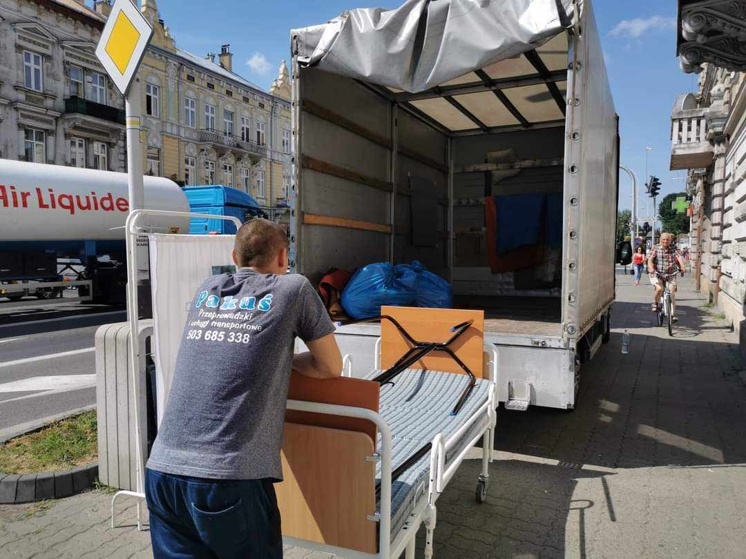 Mężczyzna w szarej koszulce z logo firmy przeprowadzkowej opiera się o wózek transportowy z łóżkiem szpitalnym na tle otwartej paki samochodu dostawczego, widoczny budynek i rowerzysta.