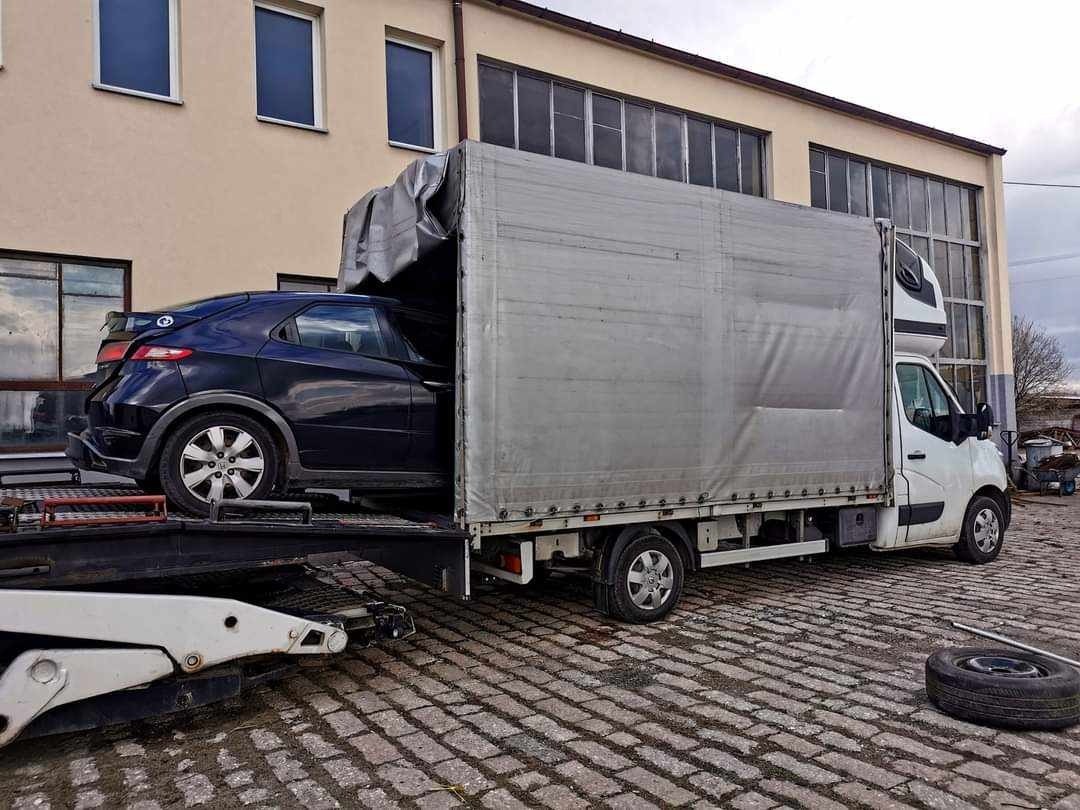 Czarny samochód osobowy Honda Civic na platformie lawety wjeżdża do białego busa z plandeką, widok z boku na tle budynku przemysłowego.