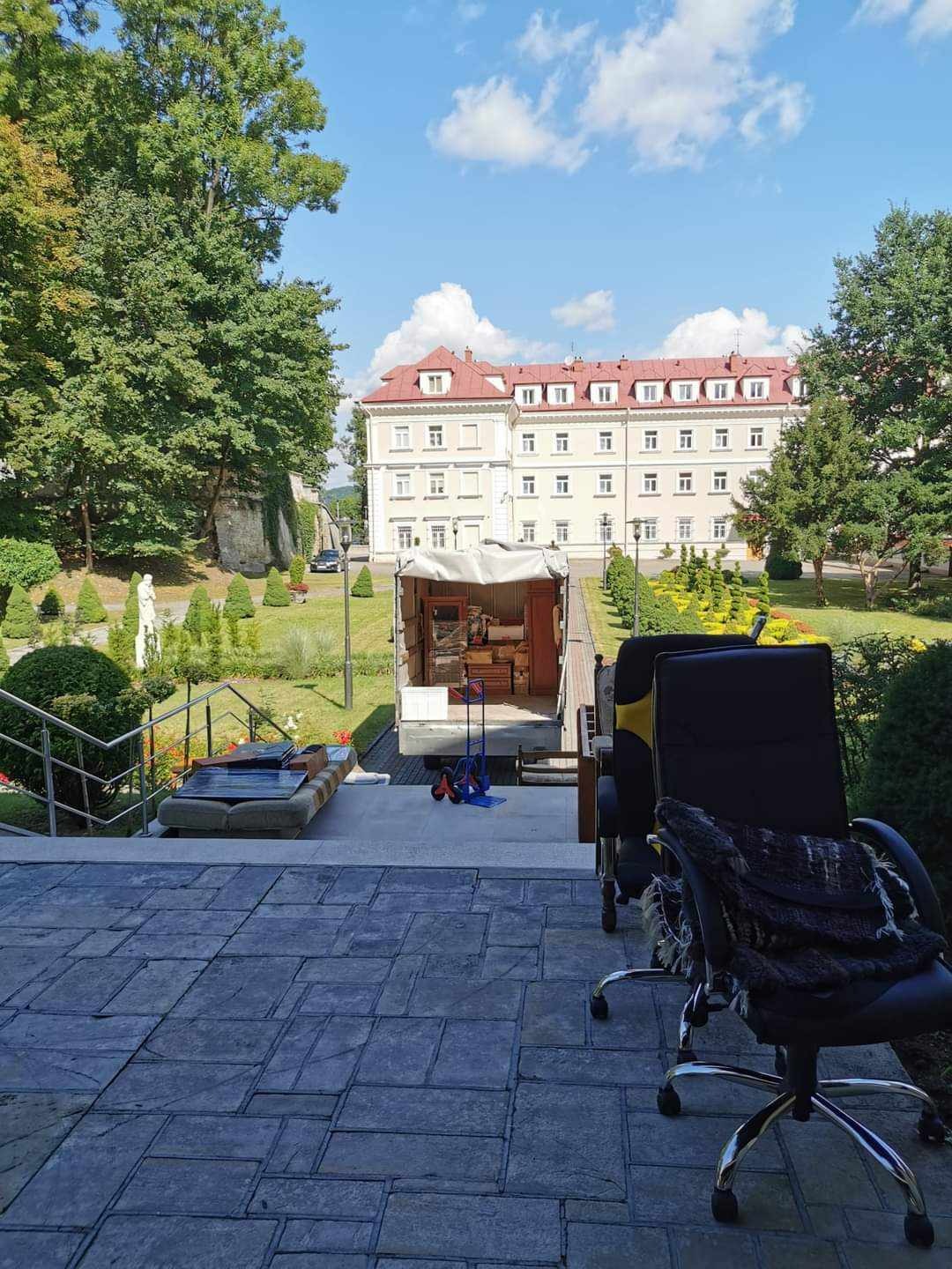 Przeprowadzka biura: meble biurowe na zewnątrz, otwarty tył furgonetki z widocznym wyposażeniem, budynek w tle, słoneczny dzień.