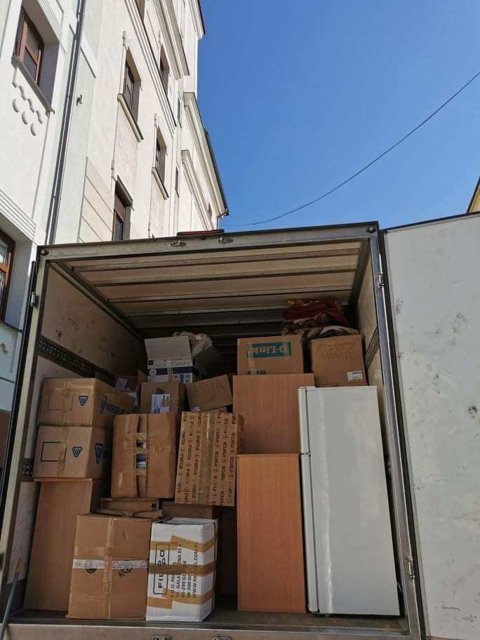 Wnętrze furgonetki wypełnione kartonami, meblami i lodówką podczas dnia przeprowadzki, z widocznym fragmentem budynku w tle.