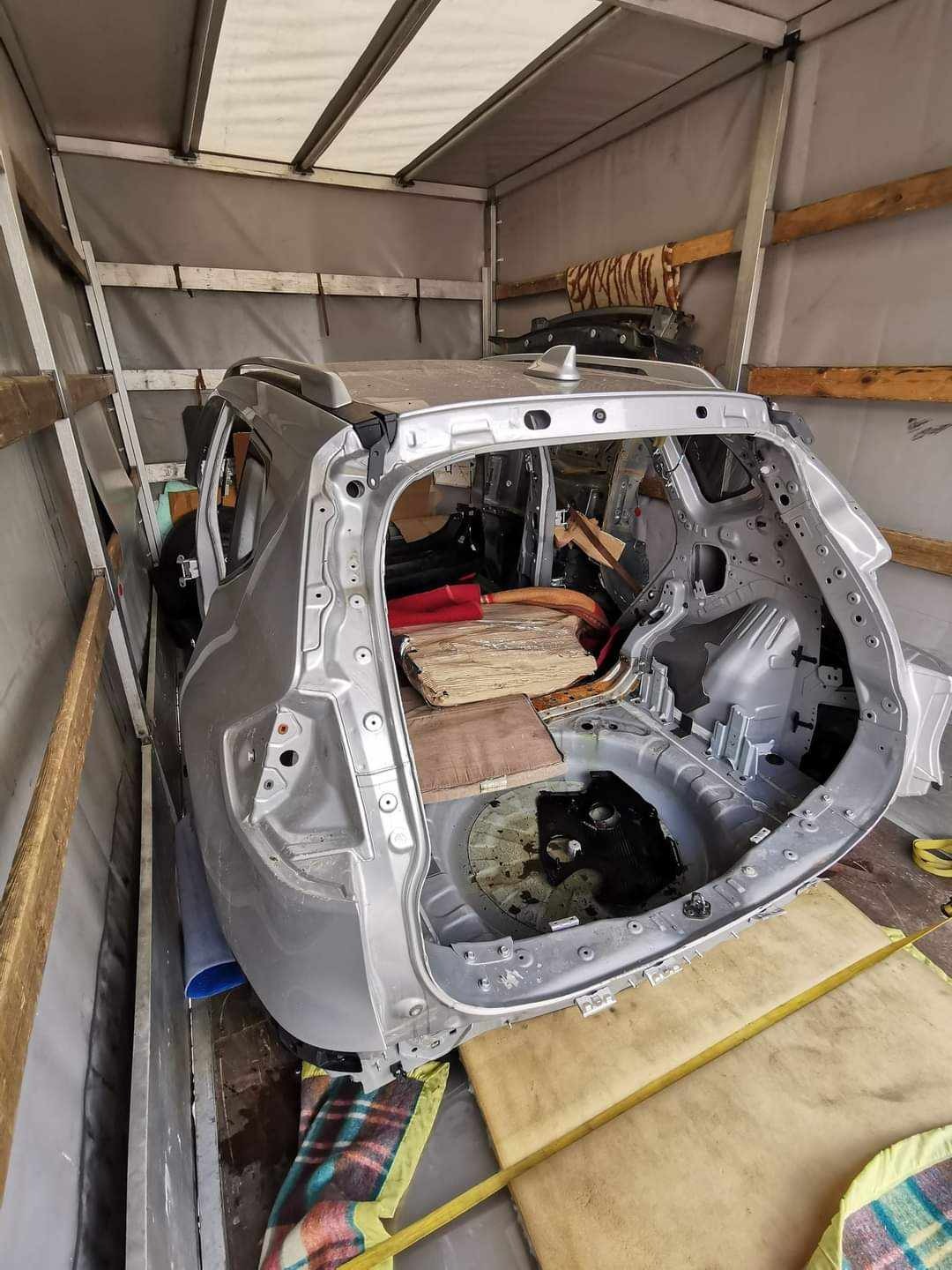 Srebrny szkielet samochodu bez karoserii, z widocznymi elementami konstrukcyjnymi, transportowany wewnątrz przestrzeni ładunkowej z drewnianymi belkami i plandeką.