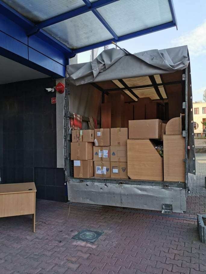 Otwarta paka ciężarówki wypełniona kartonami i meblami, częściowo przykryta szarą plandeką, zaparkowana na brukowanym placu z biurkiem obok, widoczny budynek w tle.