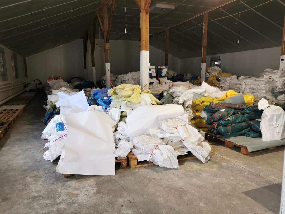 Magazyn wypełniony workami i pakunkami różnej wielkości, przygotowanymi do transportu, ułożonymi na paletach w przestronnym, ale słabo oświetlonym pomieszczeniu.