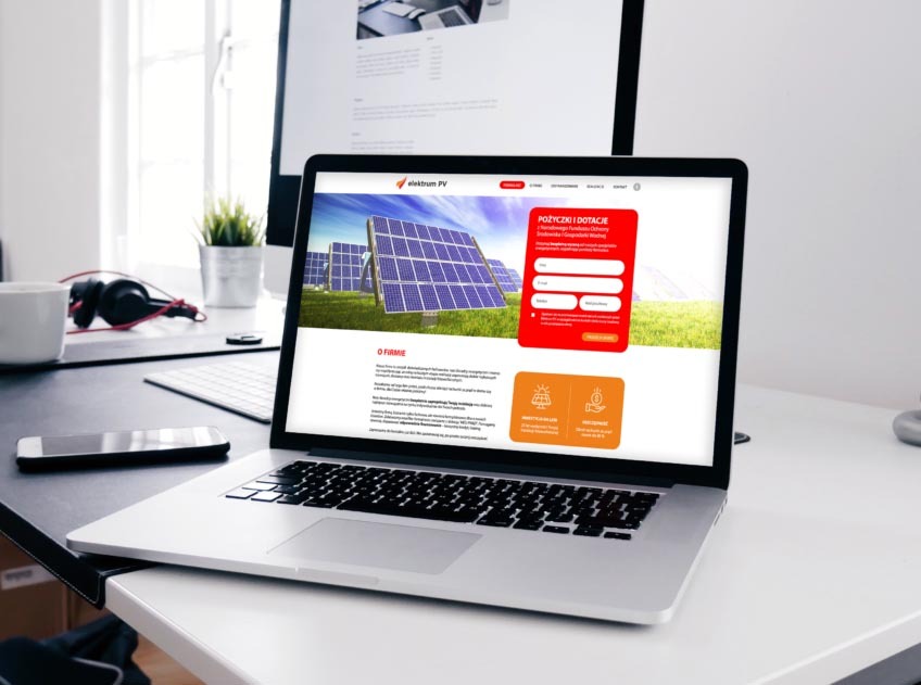 Laptop wyświetla stronę internetową firmy z panelami słonecznymi, widoczne panele słoneczne w tle strony, obok na biurku telefon i filiżanka.