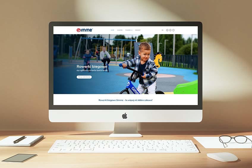 Monitor iMac wyświetlający stronę internetową z dziećmi na rowerkach biegowych marki Gimme, na biurku obok klawiatura, notatnik, tablet i okulary.