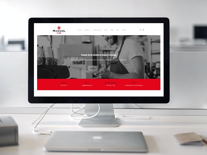 Ekran monitora wyświetlający stronę internetową kawiarni Manuel Caffe z czarno-białym zdjęciem baristy i czerwonym paskiem z danymi kontaktowymi, na biurku obok laptop i notatnik.