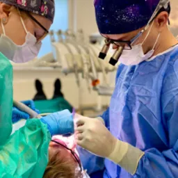 Dwóch lekarzy w uniformach i maskach, jeden z lupami, przeprowadza zabieg na pacjencie leżącym na fotelu dentystycznym, używając narzędzia stomatologicznego.