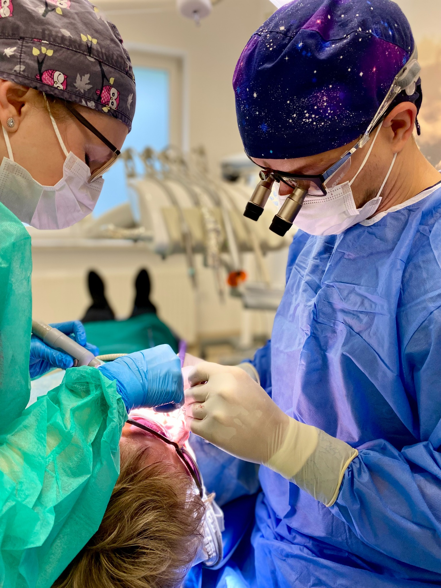 Dwóch lekarzy w uniformach i maskach, jeden z lupami, przeprowadza zabieg na pacjencie leżącym na fotelu dentystycznym, używając narzędzia stomatologicznego.