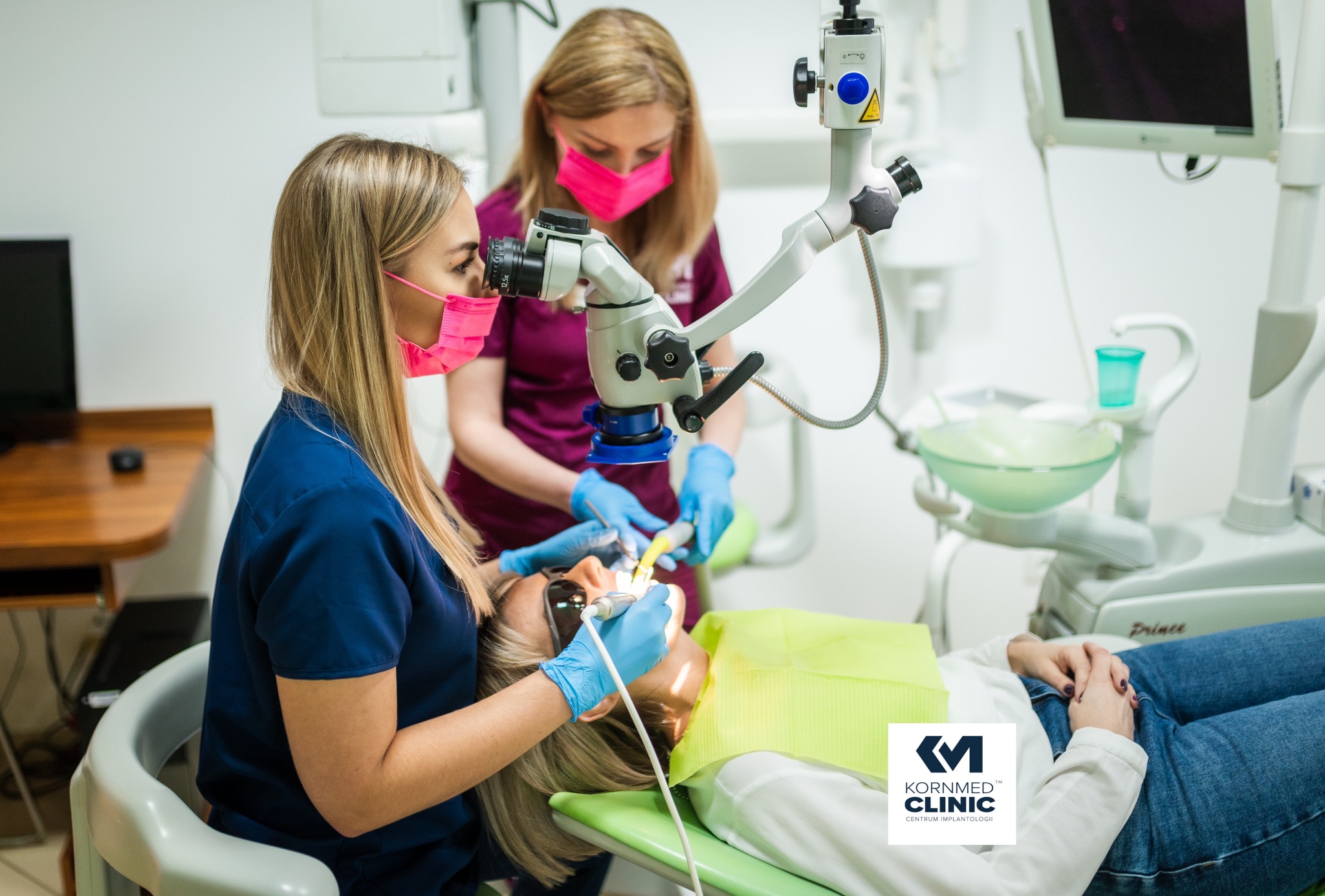 Pacjentka w okularach ochronnych podczas zabiegu stomatologicznym pod mikroskopem, wykonywanym przez dentystkę w niebieskim uniformie, asystuje druga osoba w różowej bluzie i masce, w tle...