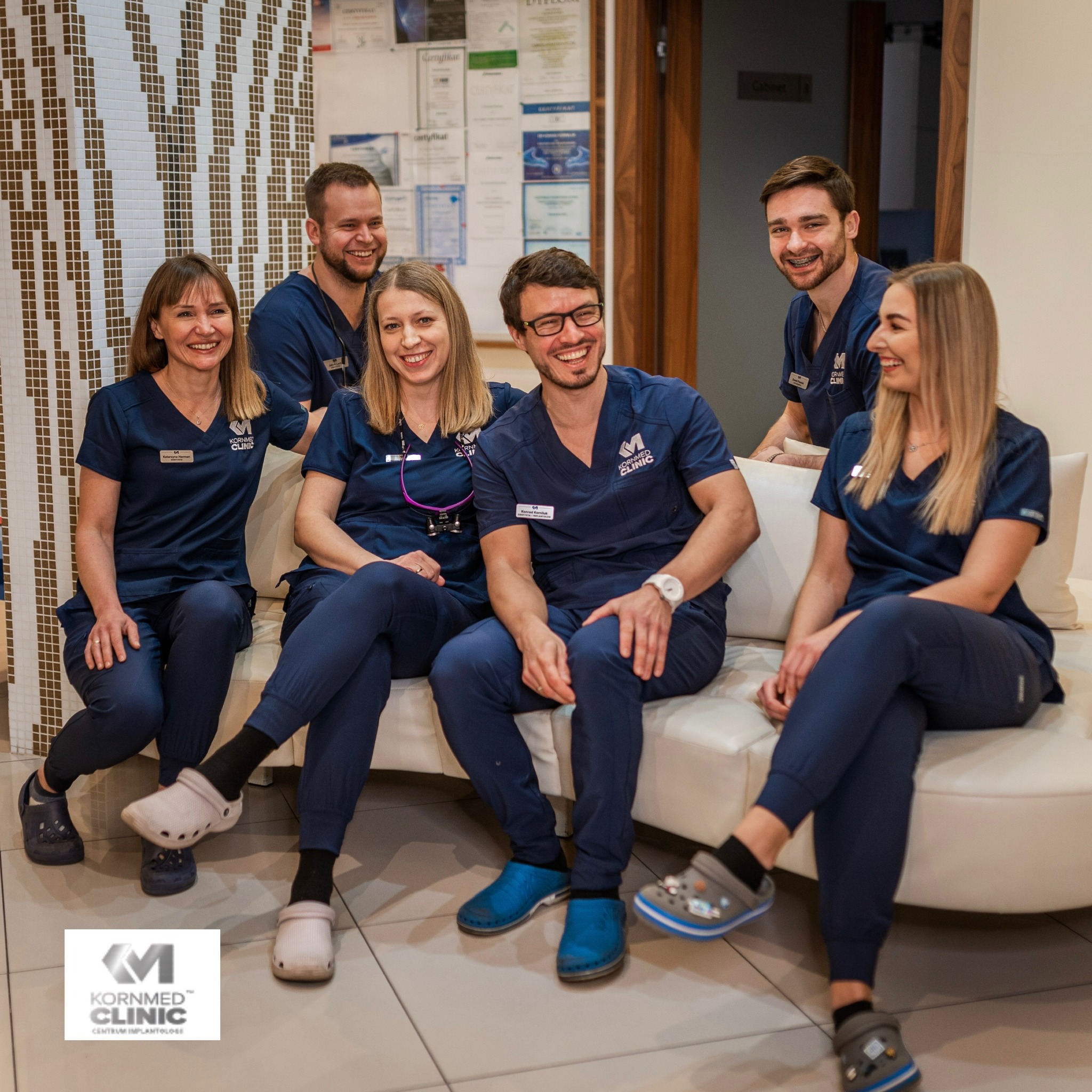 Zespół uśmiechniętych lekarzy i asystentek w granatowych uniformach Kornmed Clinic siedzi na białej sofie w nowoczesnym wnętrzu kliniki z mozaikową ścianą i certyfikatami w tle.