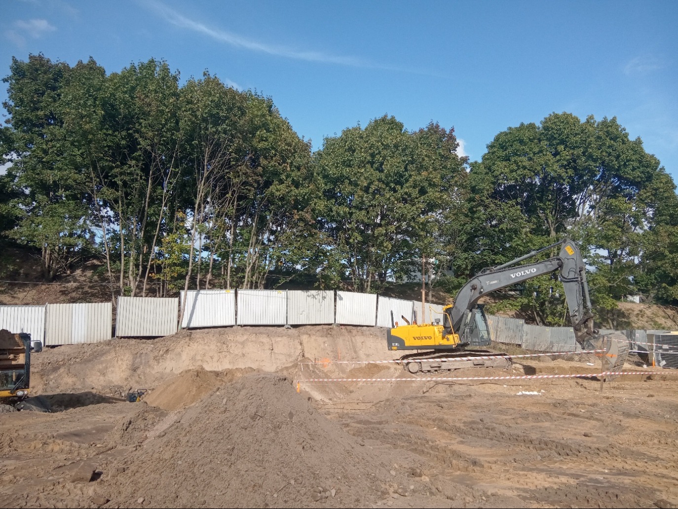 Żółta koparka Volvo na placu budowy, wykop ziemny zabezpieczony białą barierą, w tle drzewa pod błękitnym niebem.