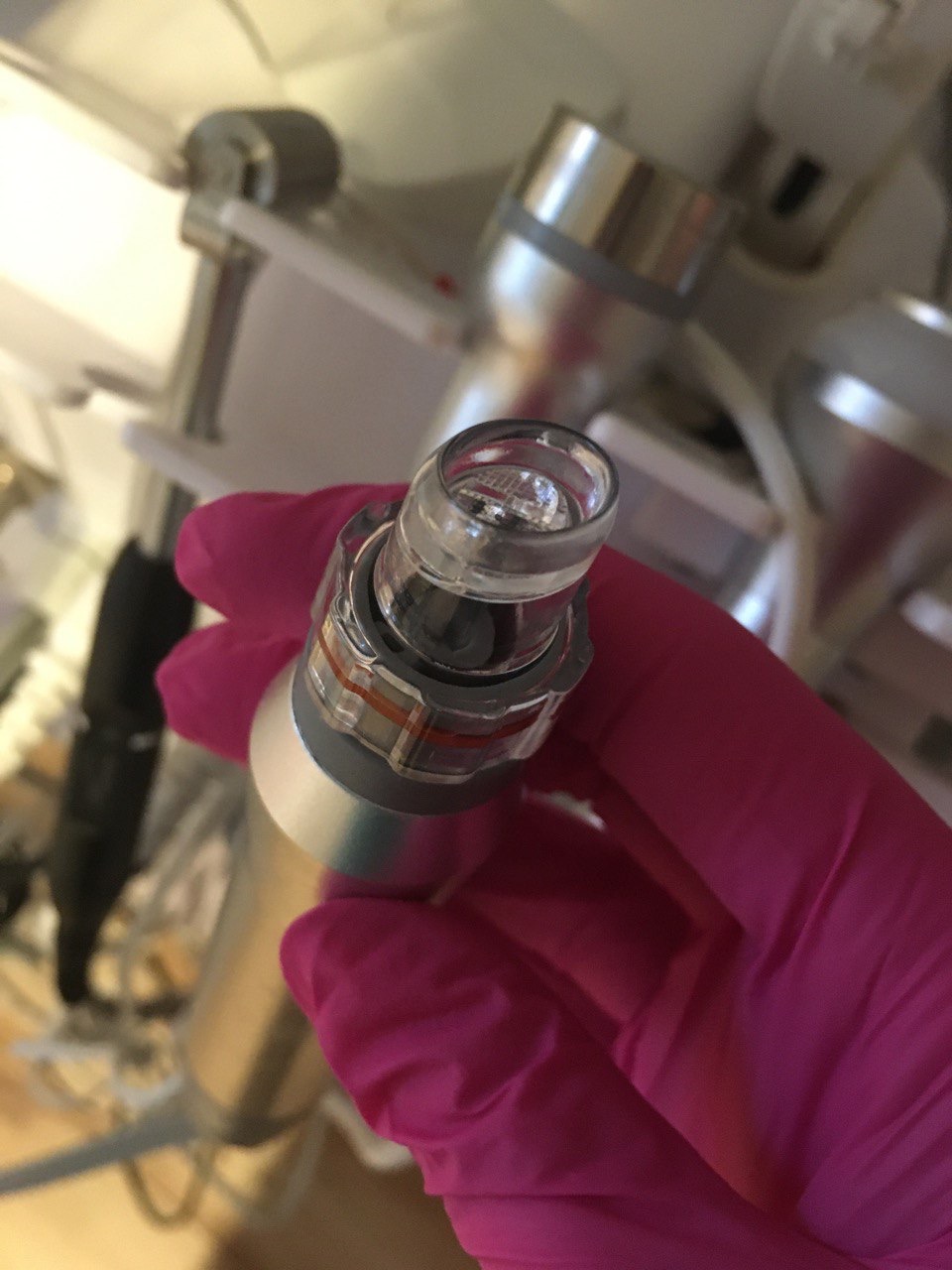 Dłoń w różowej rękawiczce trzyma końcówkę urządzenia do zabiegów kosmetycznych z przezroczystą, okrągłą końcówką, w tle widoczne inne elementy urządzenia.