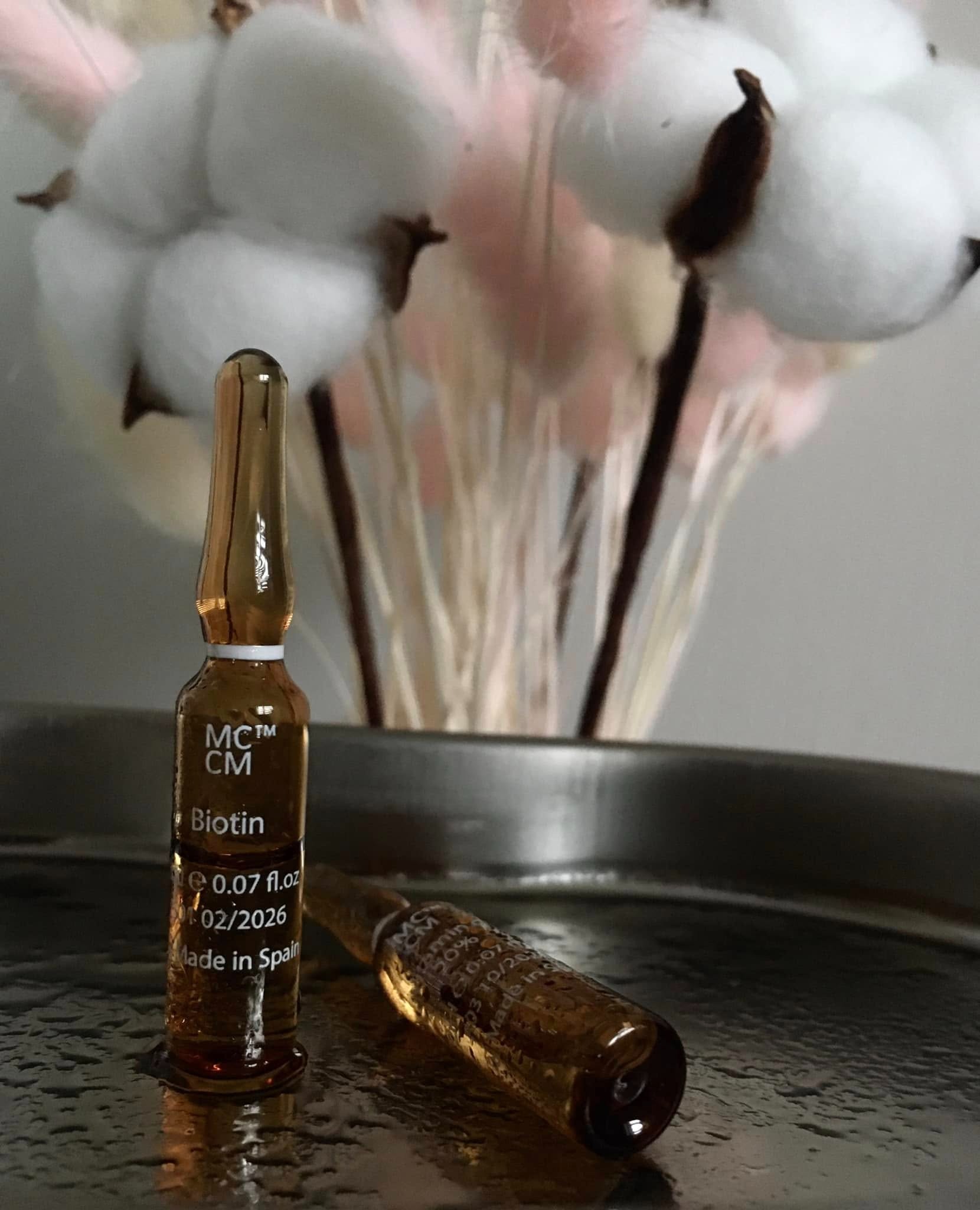 Dwie ampułki z biotyną na metalowej tacy, w tle dekoracja z bawełny. Jedna ampułka stoi, druga leży. Produkt z Hiszpanii, data ważności 02/2026.