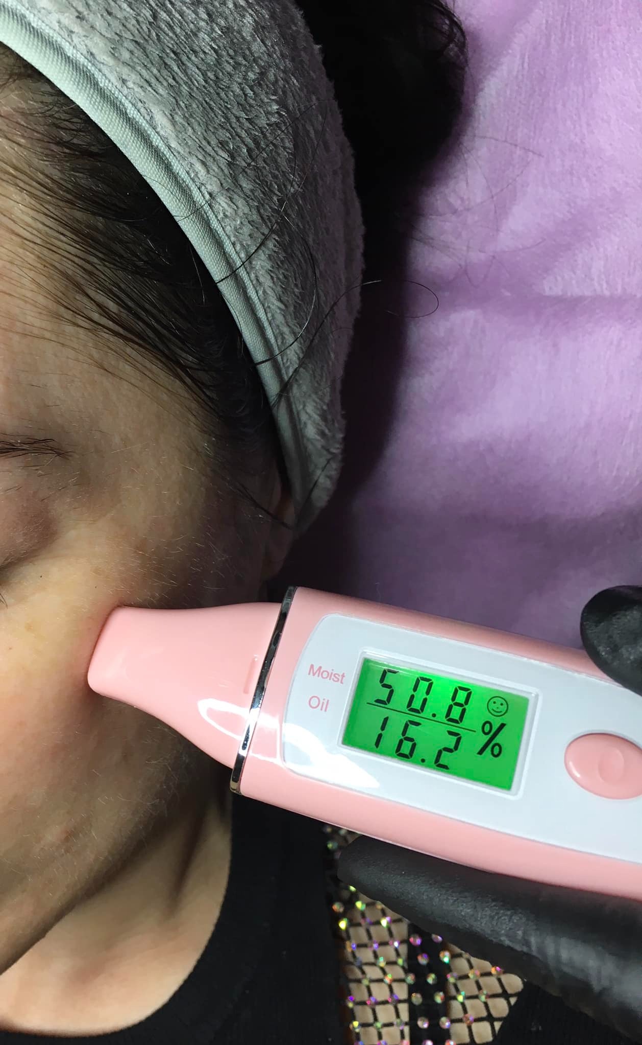 Pomiar nawilżenia skóry twarzy różowym urządzeniem diagnostycznym z wyświetlaczem, na tle fioletowej tkaniny i opaski na włosy.