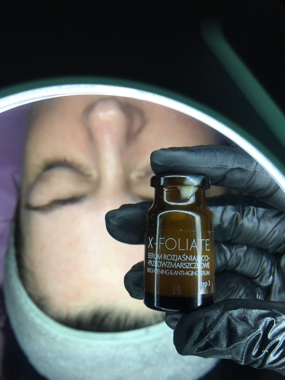Serum X-Foliate w dłoni w czarnej rękawiczce, na tle twarzy klientki pod lampą kosmetyczną. Zabieg rozjaśniająco-przeciwzmarszczkowy w salonie kosmetycznym.