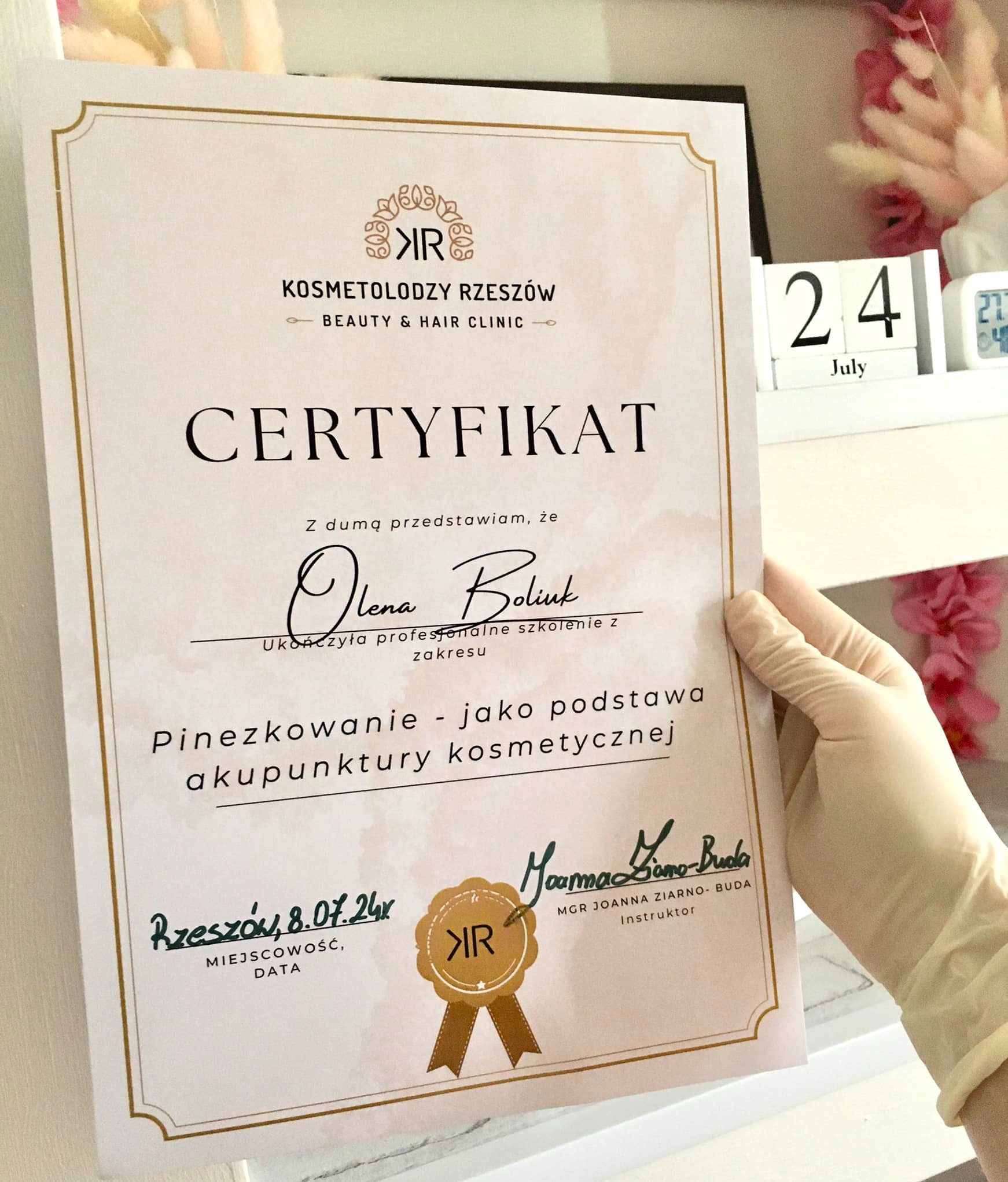 Certyfikat ukończenia szkolenia z akupunktury kosmetycznej, trzymany w dłoni w rękawiczce, z datą 8.07.24 i pieczęcią Kosmetolodzy Rzeszów.