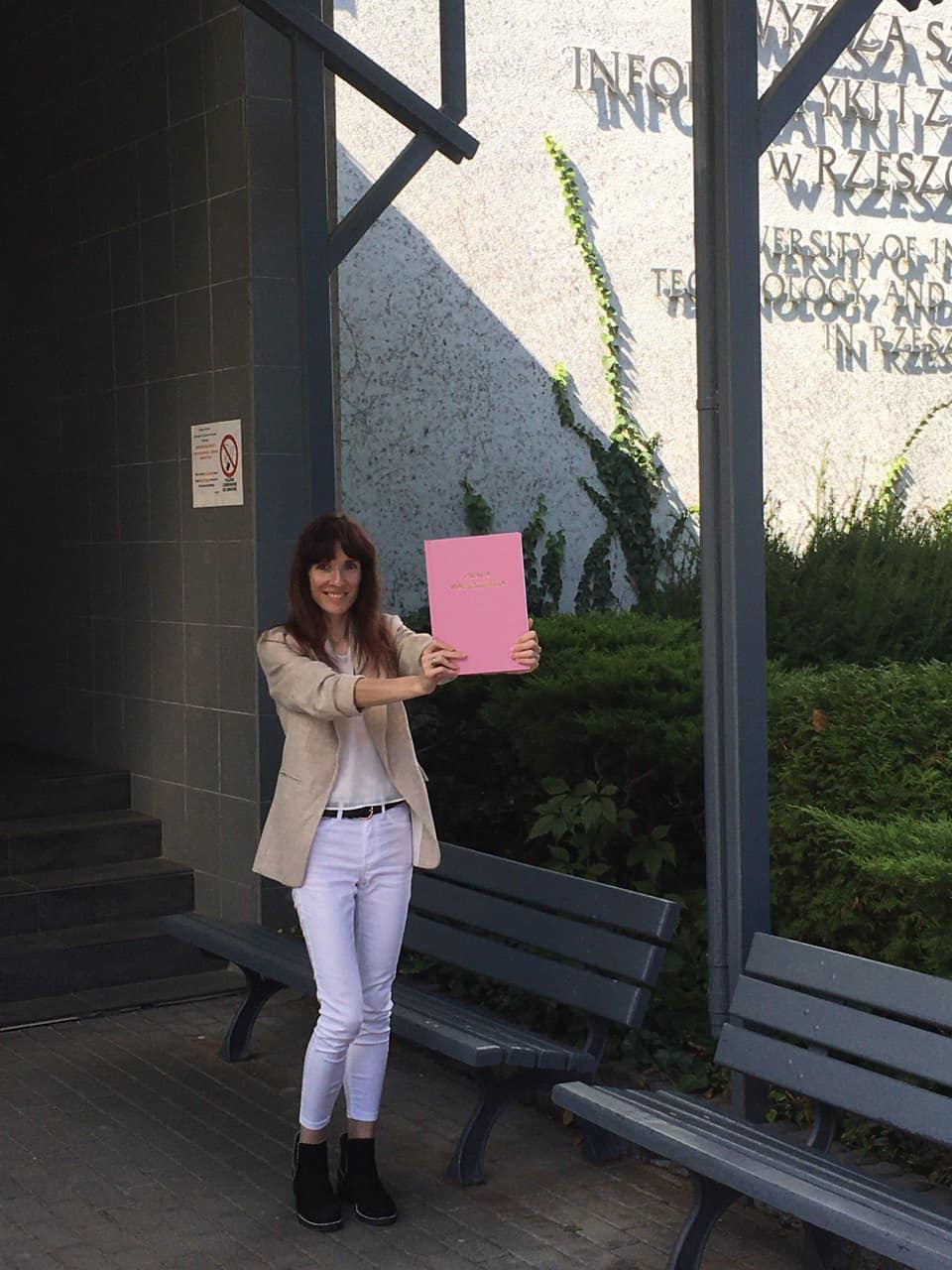 Uśmiechnięta kobieta w beżowej marynarce i białych spodniach prezentuje różowy dokument na tle budynku z napisem i zielenią.