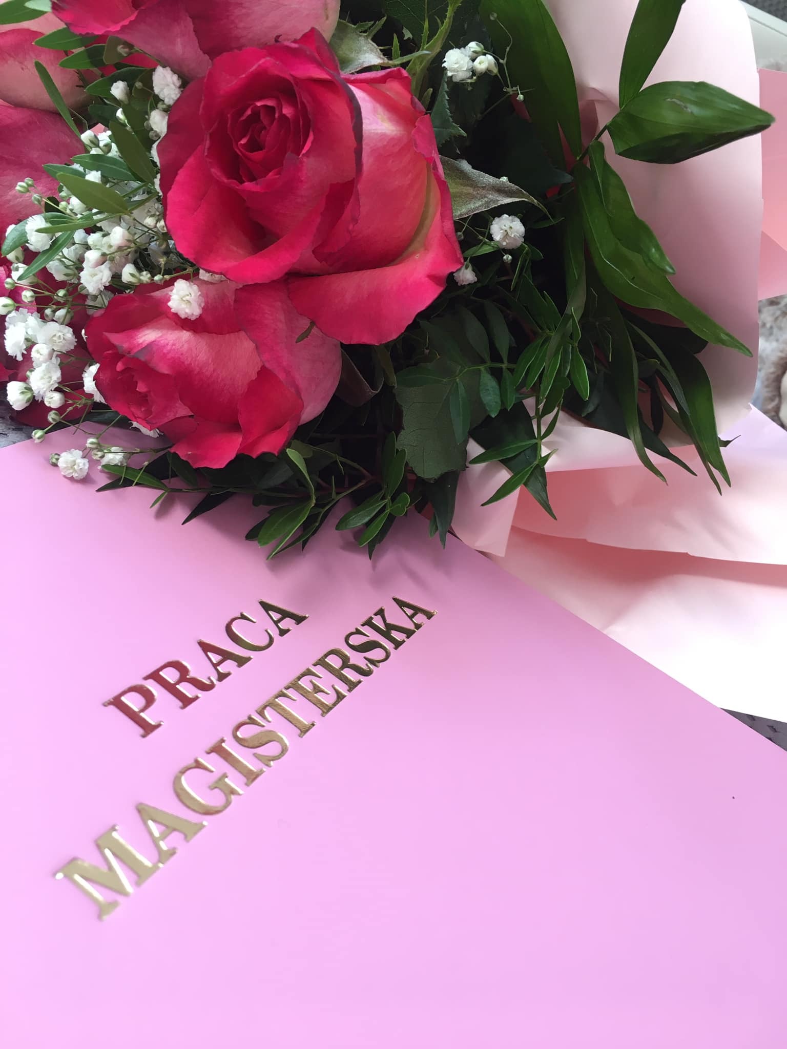 Różowy dyplom pracy magisterskiej z bukietem róż i gipsówki, złote tłoczenia na okładce, delikatne światło, ukończenie studiów.