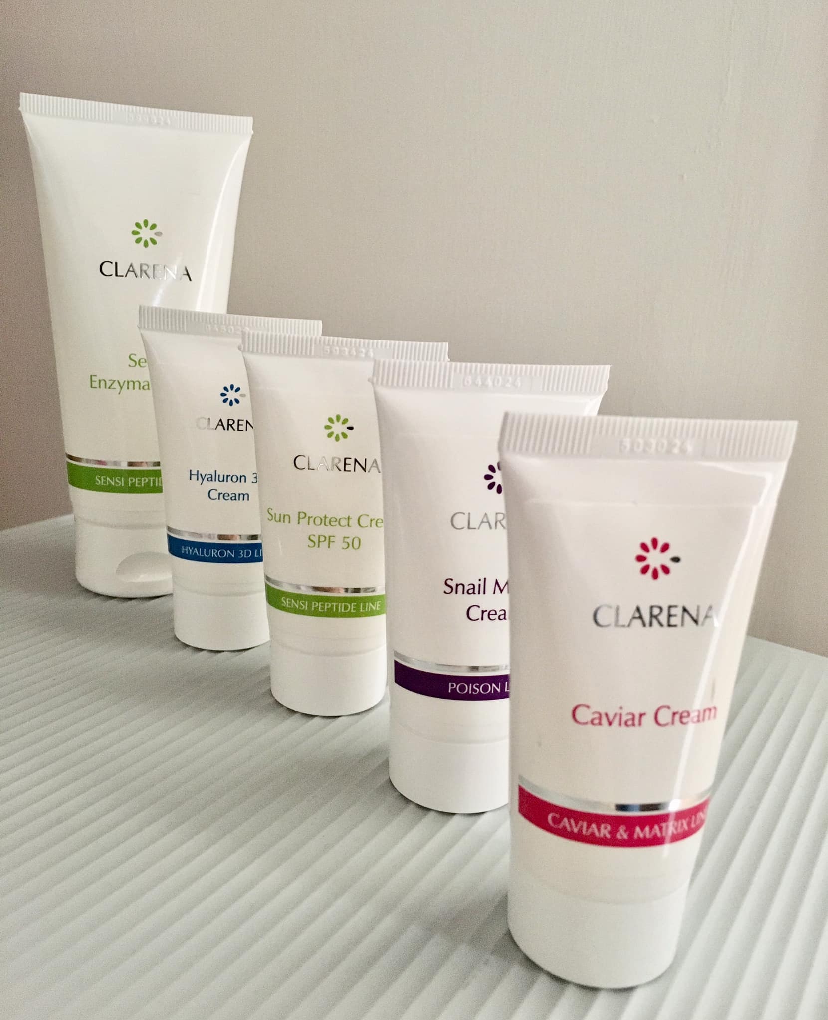 Linia kosmetyków Clarena: kremy z enzymami, kwasem hialuronowym, SPF 50, śluzem ślimaka i kawiorem. Produkty ułożone w rzędzie na teksturowanym tle.
