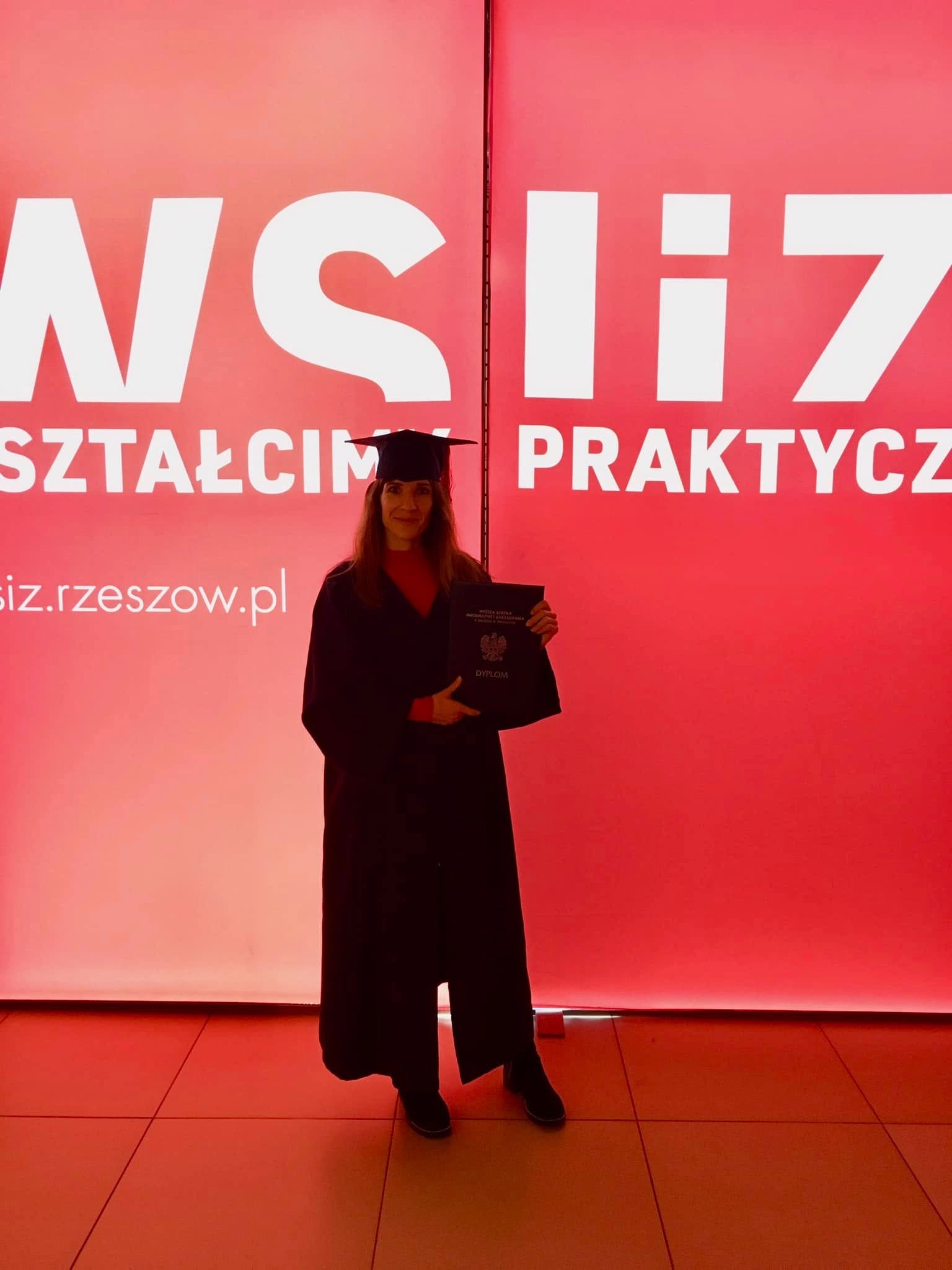 Uśmiechnięta absolwentka w todze i birecie trzyma dyplom na tle czerwonej ściany z logo uczelni WSIiZ. Widoczne całe ciało, oświetlenie studyjne.