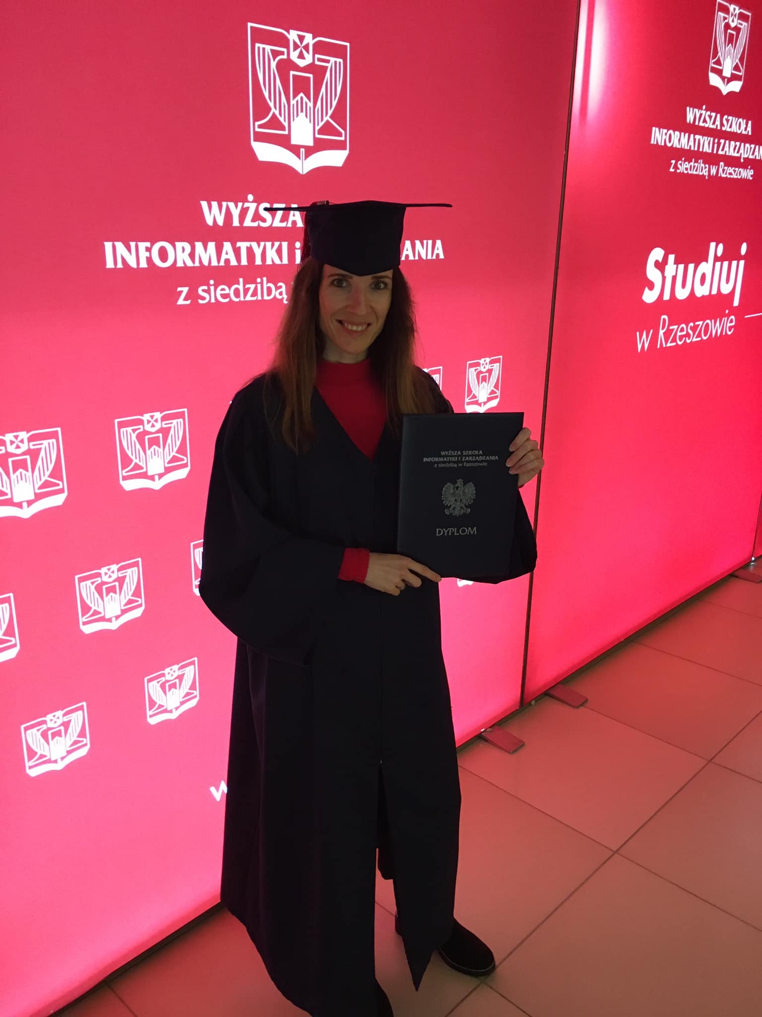 Absolwentka w todze i birecie, uśmiechnięta, trzyma dyplom na tle czerwonej ścianki z logo uczelni. Widoczny napis: Wyższa Szkoła Informatyki i Zarządzania.