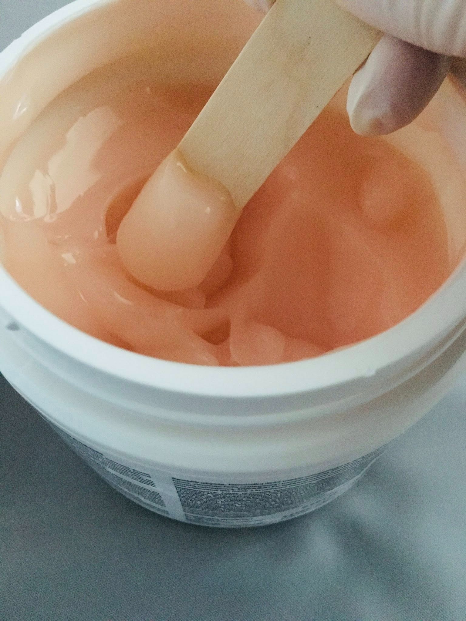 Różowa pasta cukrowa w białym pojemniku, nabierana drewnianą szpatułką przez osobę w rękawiczce. Przygotowanie do depilacji w salonie.