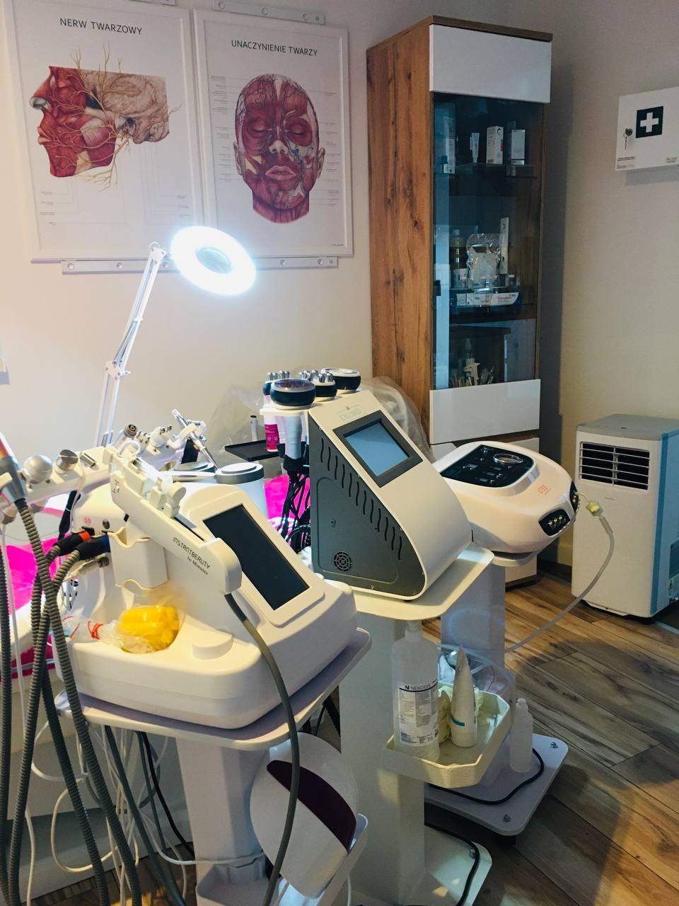 Salon kosmetyczny: widok na nowoczesne urządzenia do zabiegów na ciało, plakaty anatomiczne na ścianie i witryna z kosmetykami w tle.