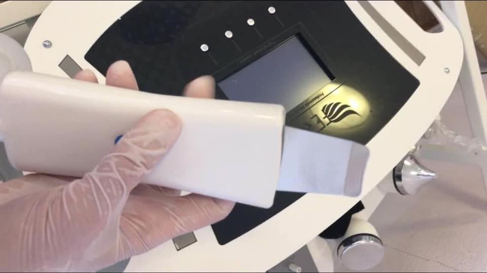 Dłoń w rękawiczce trzyma białą głowicę urządzenia do peelingu kawitacyjnego na tle panelu sterowania urządzenia kosmetycznego.