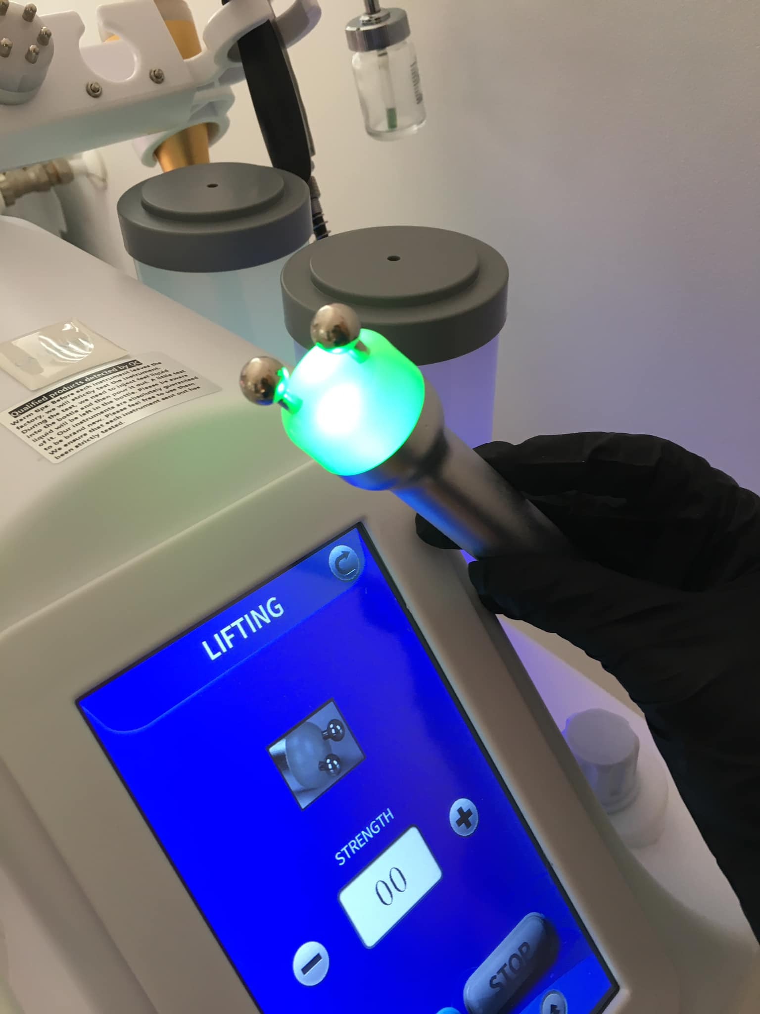 Urządzenie do liftingu twarzy z zielonym światłem, obsługiwane przez osobę w czarnych rękawiczkach ochronnych, widoczny ekran dotykowy z napisem 'Lifting' i ustawieniem 'Strength 00'.