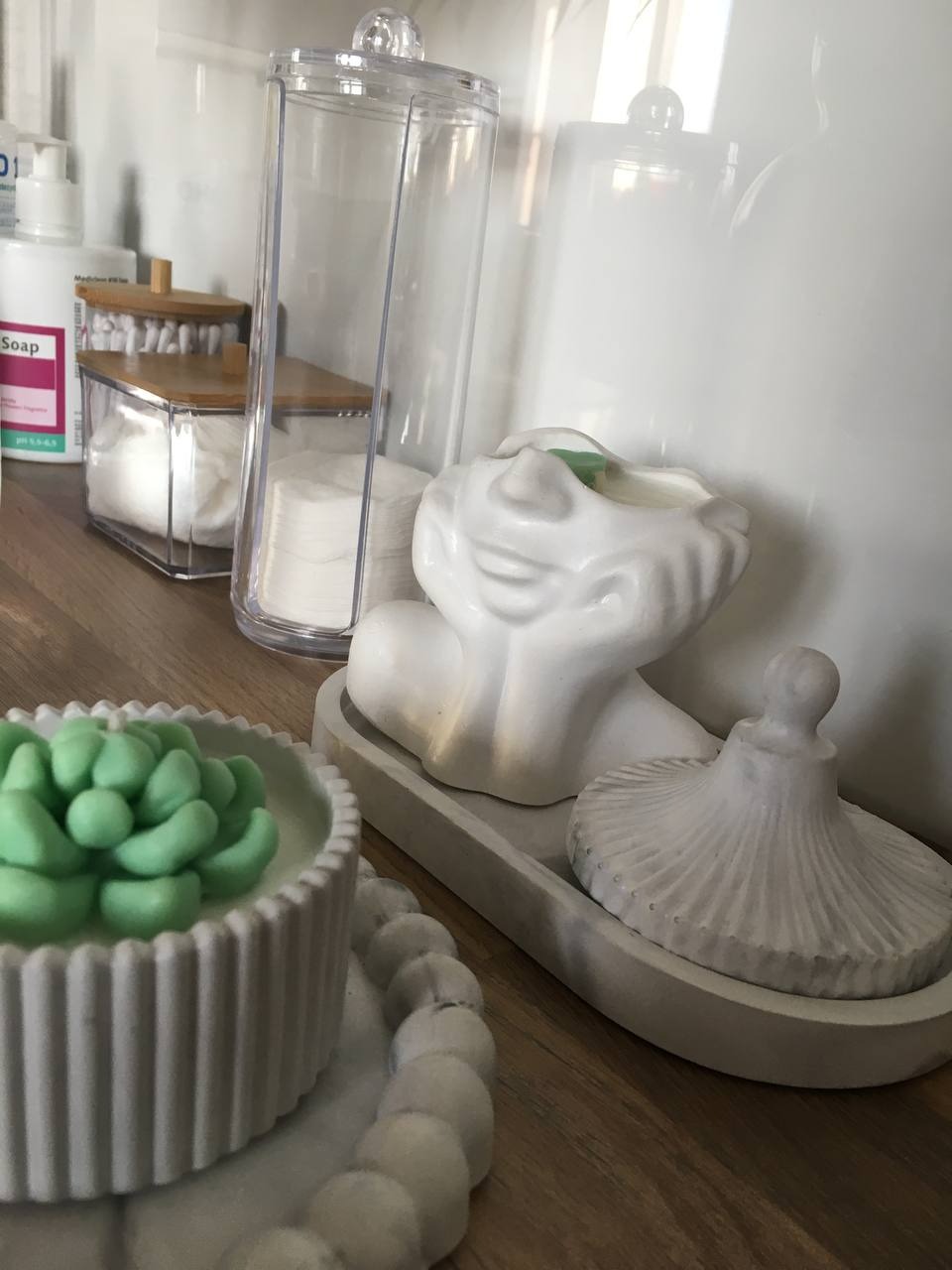 Dekoracyjny zestaw kosmetyczny w odcieniach bieli i szarości: pojemniki na waciki, ceramiczna figurka głowy kobiety z zielonym akcentem oraz pędzel do pudru na drewnianej półce.