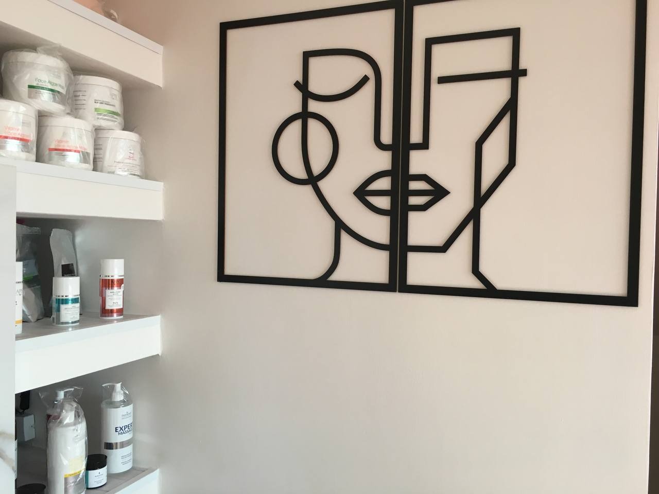 Wnętrze salonu kosmetycznego z minimalistyczną, dwuczęściową dekoracją ścienną przedstawiającą abstrakcyjną twarz oraz półkami z kosmetykami i preparatami.