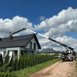 Czarny dźwig teleskopowy K100 RHX z pracownikiem w koszu na dachu domu z panelami słonecznymi, błękitne niebo z cumulusami w tle.