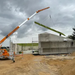 Pomarańczowy dźwig podnosi element konstrukcyjny na budowę hali szkieletowej, widoczne rusztowania i pracownicy na wysokości.