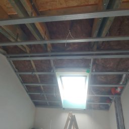 Adam Kawończyk BRAT-BUD-WYKOŃCZENIA - Konstrukcja metalowa sufitu podwieszanego z oknem dachowym, ocieplona wełną mineralną, widoczna z dołu, drabina w tle.