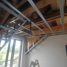 Adam Kawończyk BRAT-BUD-WYKOŃCZENIA - Montaż stelaża z profili stalowych pod płytę kartonowo-gipsową na poddaszu, widoczne ocieplenie wełną mineralną między krokwiami, okno dachowe z widokiem na zieleń, drabina w rogu pomieszczenia.