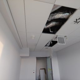 Adam Kawończyk BRAT-BUD-WYKOŃCZENIA - Biały sufit podwieszany z widocznymi elementami instalacji wentylacyjnej i klimatyzacji w otworach, fragment pustego, jasnego pomieszczenia z drzwiami w tle.