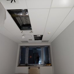 Adam Kawończyk BRAT-BUD-WYKOŃCZENIA - Montaż sufitu podwieszanego z widocznymi otworami rewizyjnymi i instalacją wentylacyjną, częściowo zakryty folią ochronną.