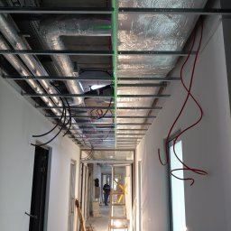 Adam Kawończyk BRAT-BUD-WYKOŃCZENIA - Korytarz w trakcie prac wykończeniowych, widoczne instalacje pod sufitem, przewody elektryczne, drabina i pracownik w oddali.