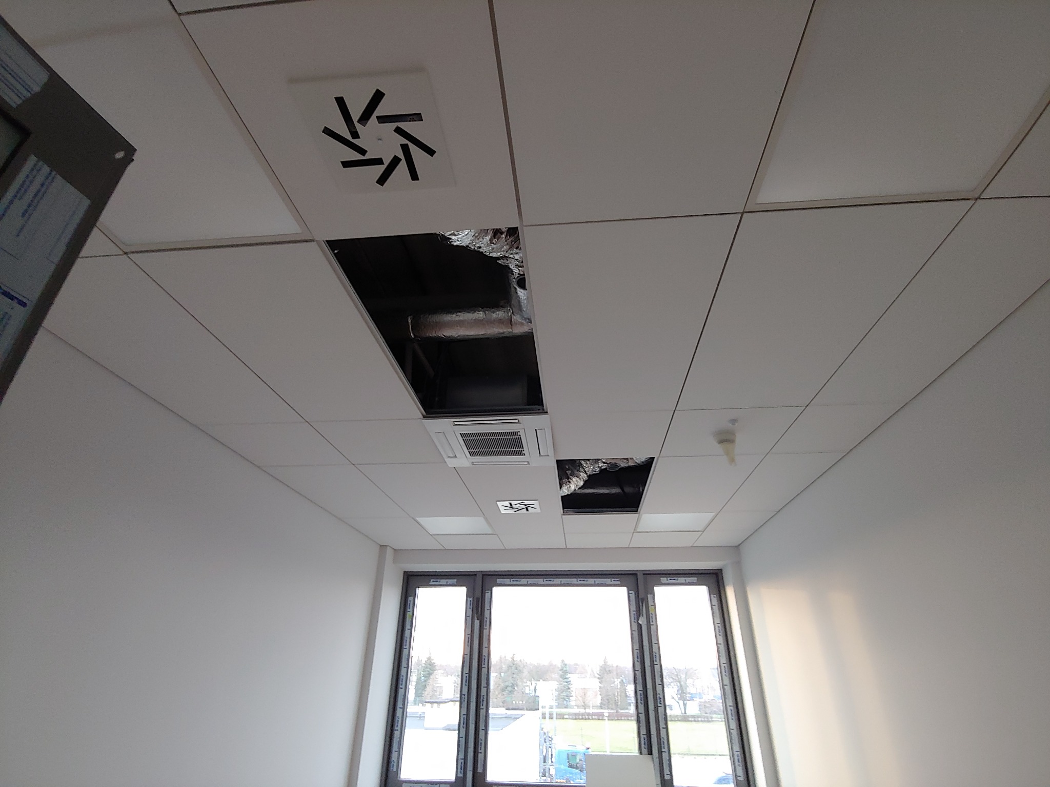 Montaż sufitu podwieszanego kasetonowego z widocznymi otworami na instalacje wentylacyjne i klimatyzacyjne, fragmenty instalacji widoczne w otworach, białe ściany i okna w tle.