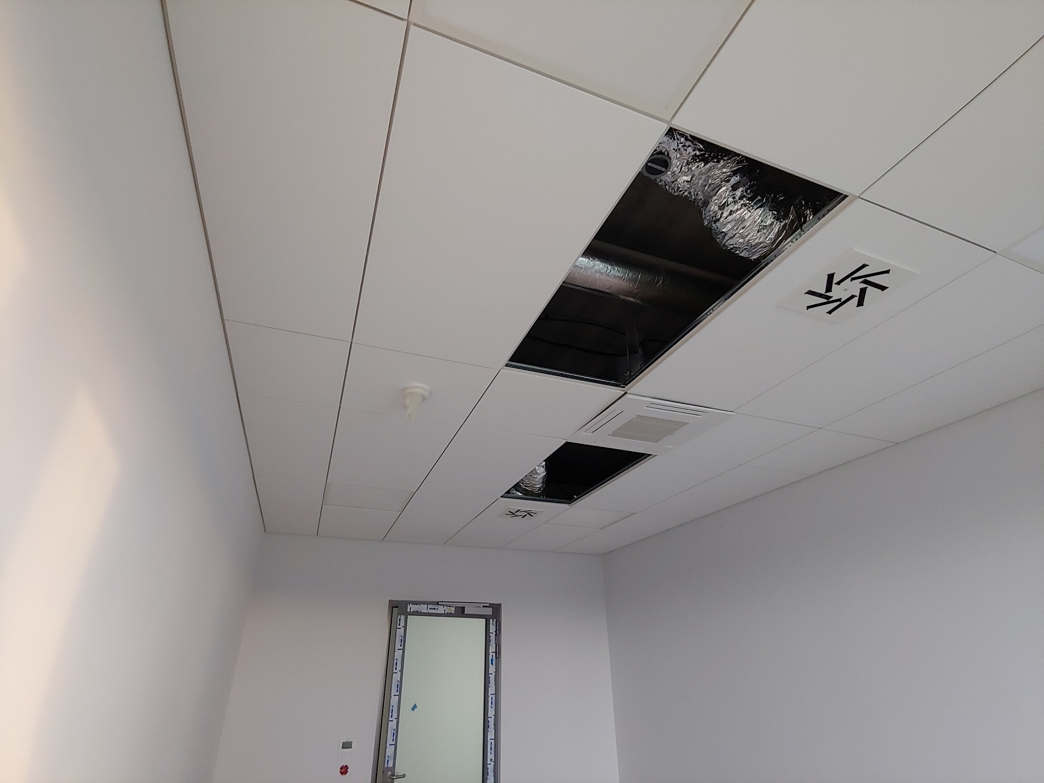 Biały sufit kasetonowy z widocznymi otworami na instalacje, w tle białe ściany i drzwi.