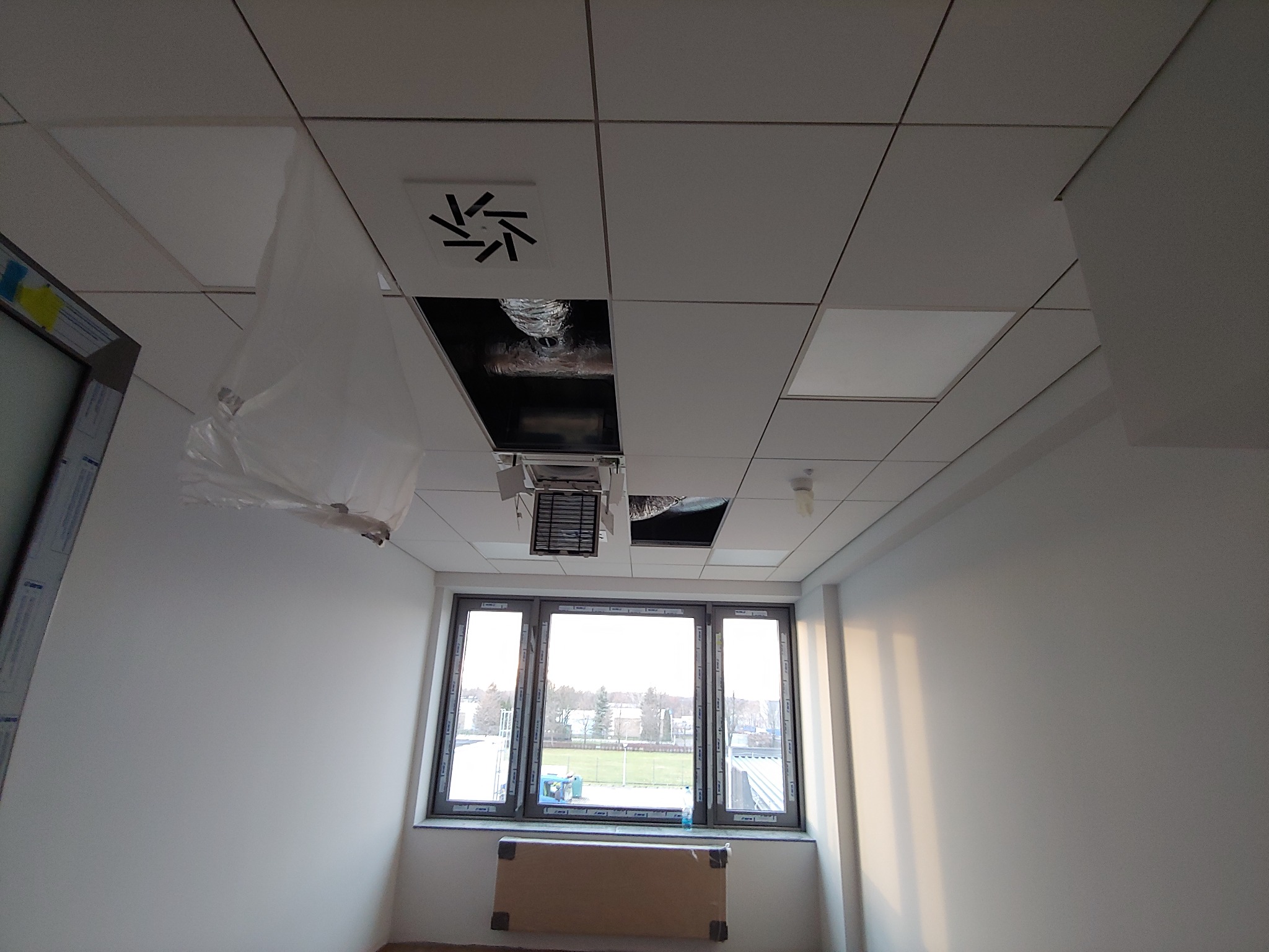 Wnętrze biurowe w trakcie wykańczania: biały sufit podwieszany z otworami na instalacje, widoczne przewody wentylacyjne i klimatyzacja, ściany pomalowane na biało, duże okno z widokiem na zewnątrz,...