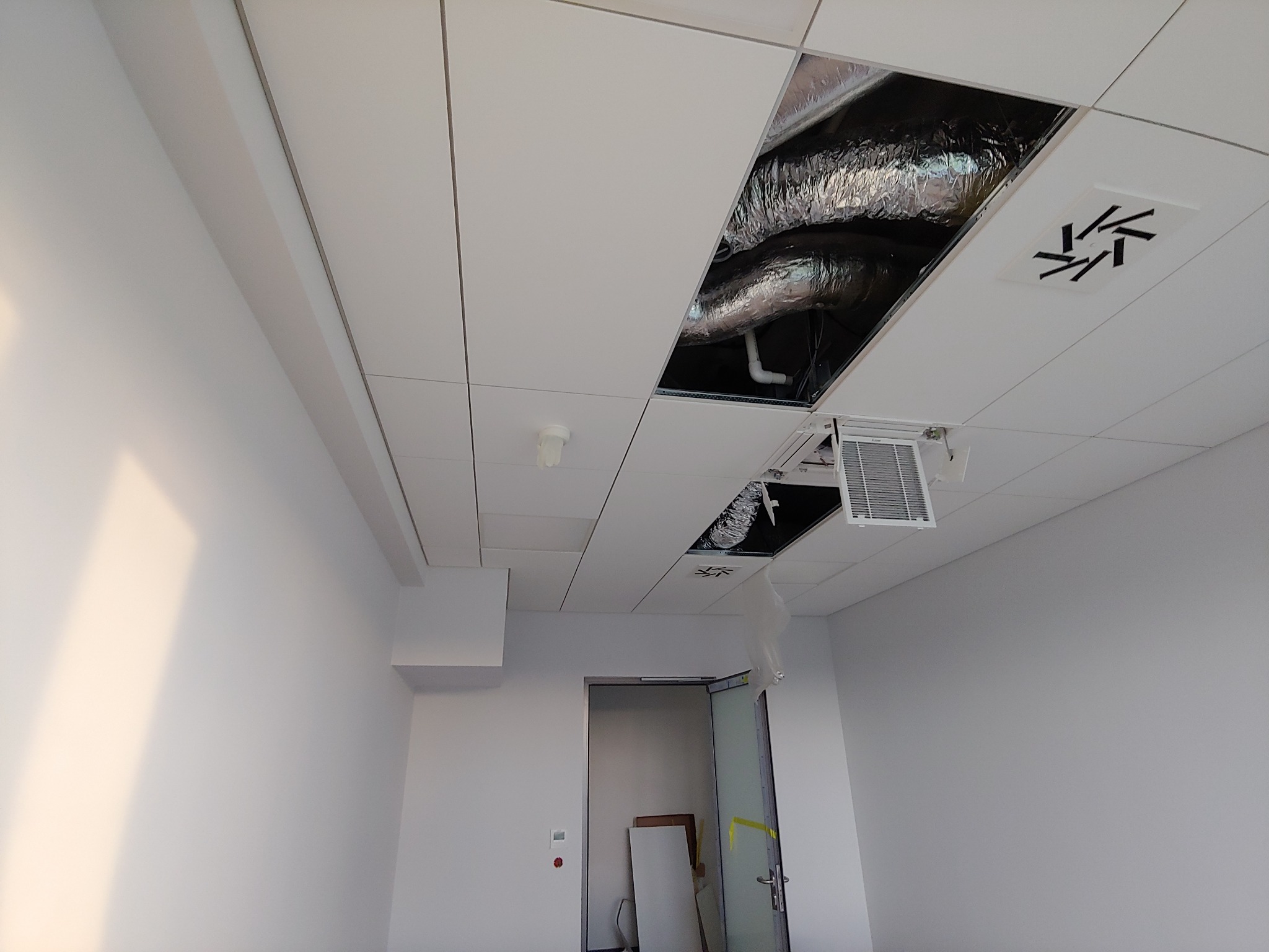 Biały sufit podwieszany z widocznymi elementami instalacji wentylacyjnej i klimatyzacji w otworach, fragment pustego, jasnego pomieszczenia z drzwiami w tle.