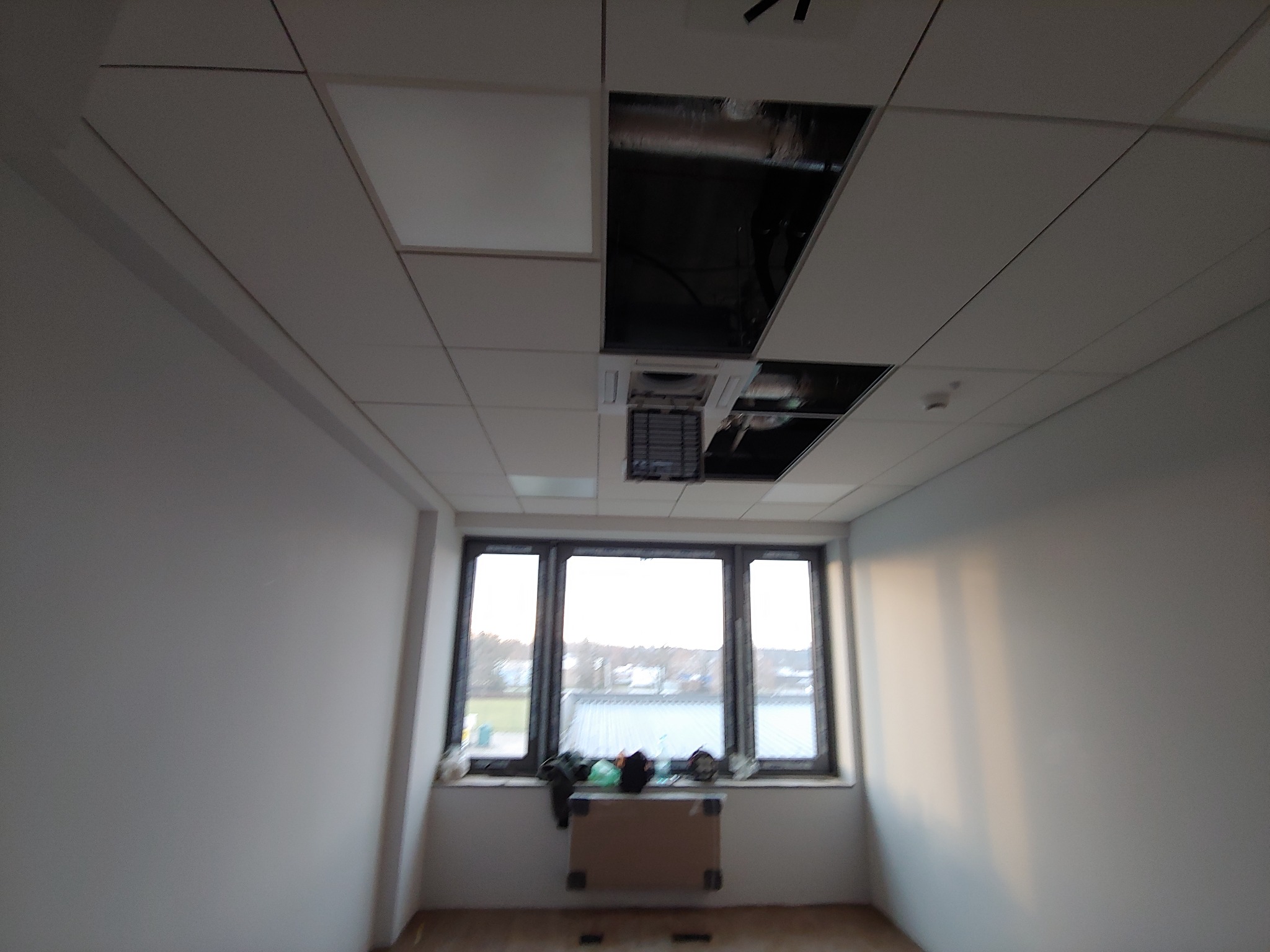 Wnętrze biura w trakcie montażu podwieszanego sufitu kasetonowego, widoczne otwarte przestrzenie na instalacje i wentylację, ściany przygotowane do malowania, okno z widokiem na zewnątrz.