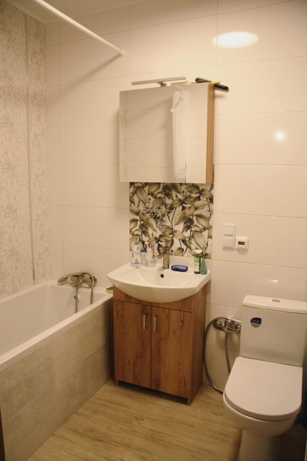 Jasna łazienka w Gdańsku po remoncie, z wanną, umywalką z szafką w kolorze drewna i toaletą; na ścianie nad umywalką dekoracyjne płytki z motywem liści.