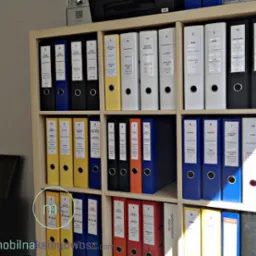 P&oacute;łka regału wypełniona kolorowymi segregatorami z etykietami, zawierającymi dokumenty z r&oacute;żnych lat. Widoczne segregatory w kolorach: niebieskim, czarnym, ż&oacute;łtym, białym i pomarańczowym.