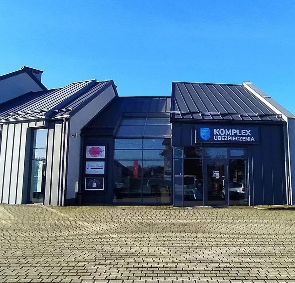 Budynek biura ubezpieczeniowego 'Komplex Ubezpieczenia' z szarymi ścianami, dużymi oknami i logo firmy nad wejściem, widziany z poziomu brukowanego chodnika.